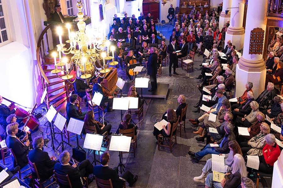 Overzicht van de kerk tijdens de uitvoering: het publiek rechts met tekstboeken op schoot, het orkest links, en een brandende kroonluchter boven de zaal.